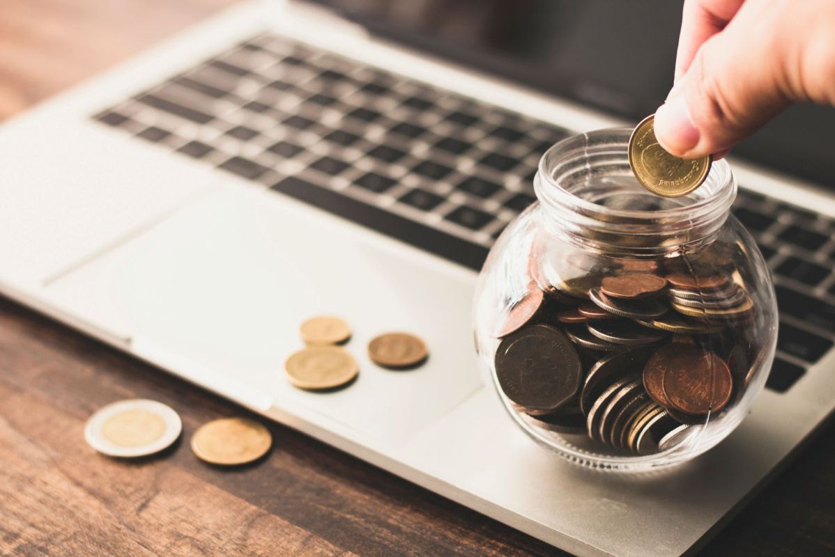 כמה עולה פרסום ממומן בגוגל. כף יד מכניסה מטבע לצנצנת מלאה מטבעות שיושבת על מקלדת לפטופ.